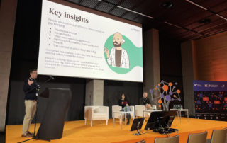 A photo of three people on a dais: a man behind a lecturn and a man and a woman seated on chairs to his left. Behind them is a presentation screen with the title “Key insights” and a drawing of a balding man with a beard pointing to his right.