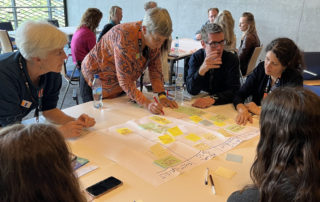 Several people around a table on which is an A0 sheet with sticky notes on it. A women is standing and writing something on the document.